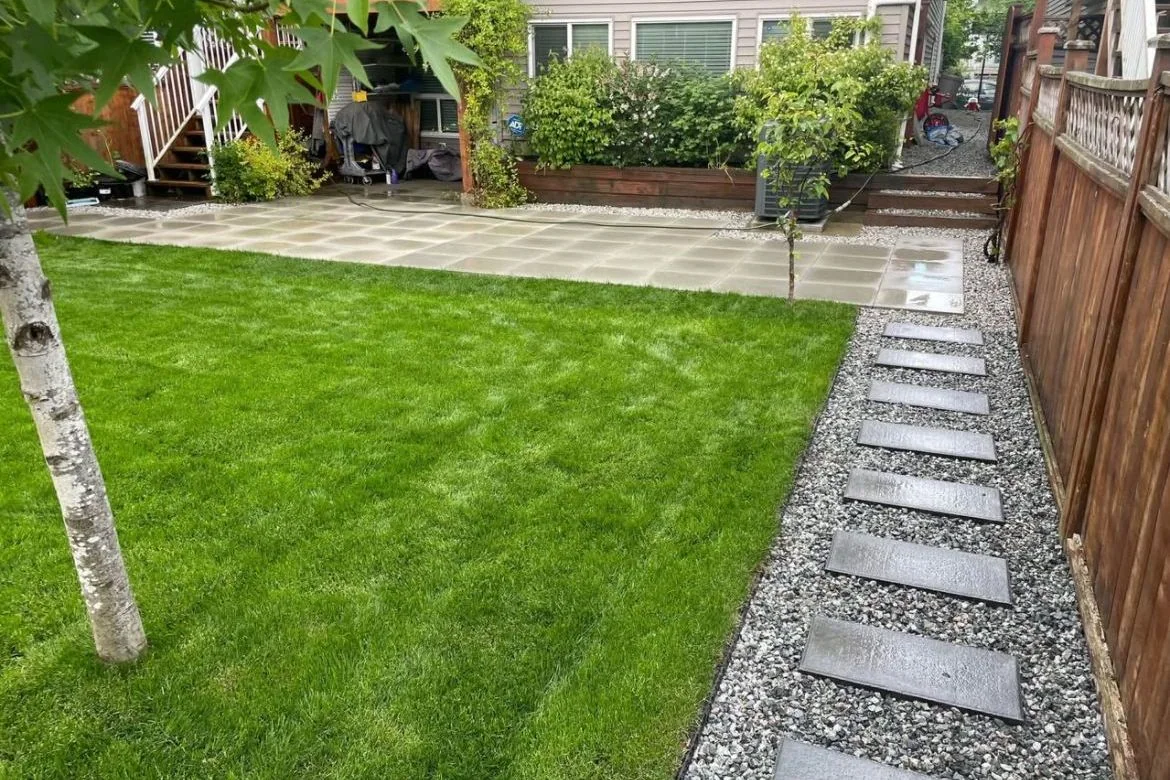 Green backyard lawn framed by gravel paths, trees, and a concrete patio